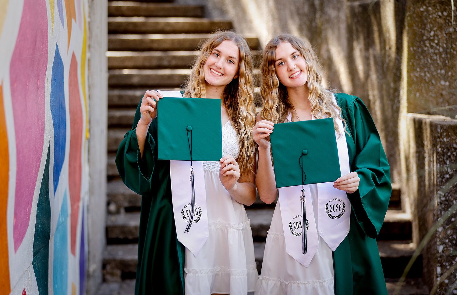 Two happy graduates in green caps and gowns proudly display diplomas near a colorful painted mural.