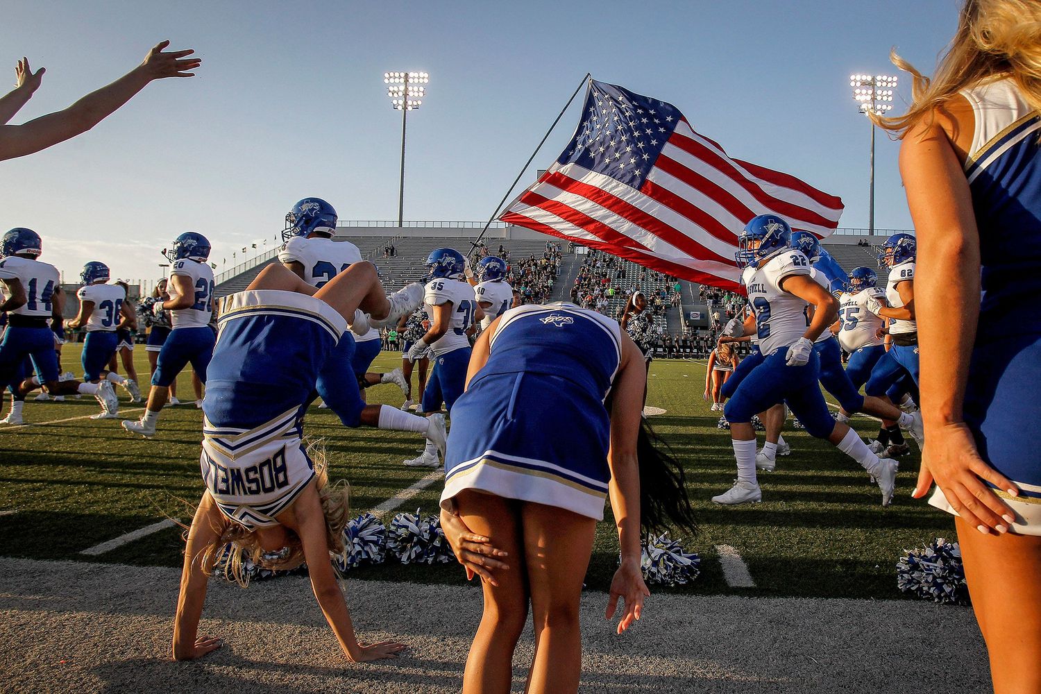 Cheerleaders hold American flag as football team runs onto field.