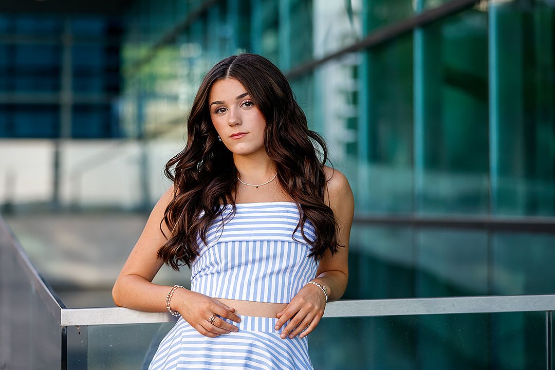 Young woman in striped blue dress poses gracefully outdoors near glass building with long wavy dark hair.