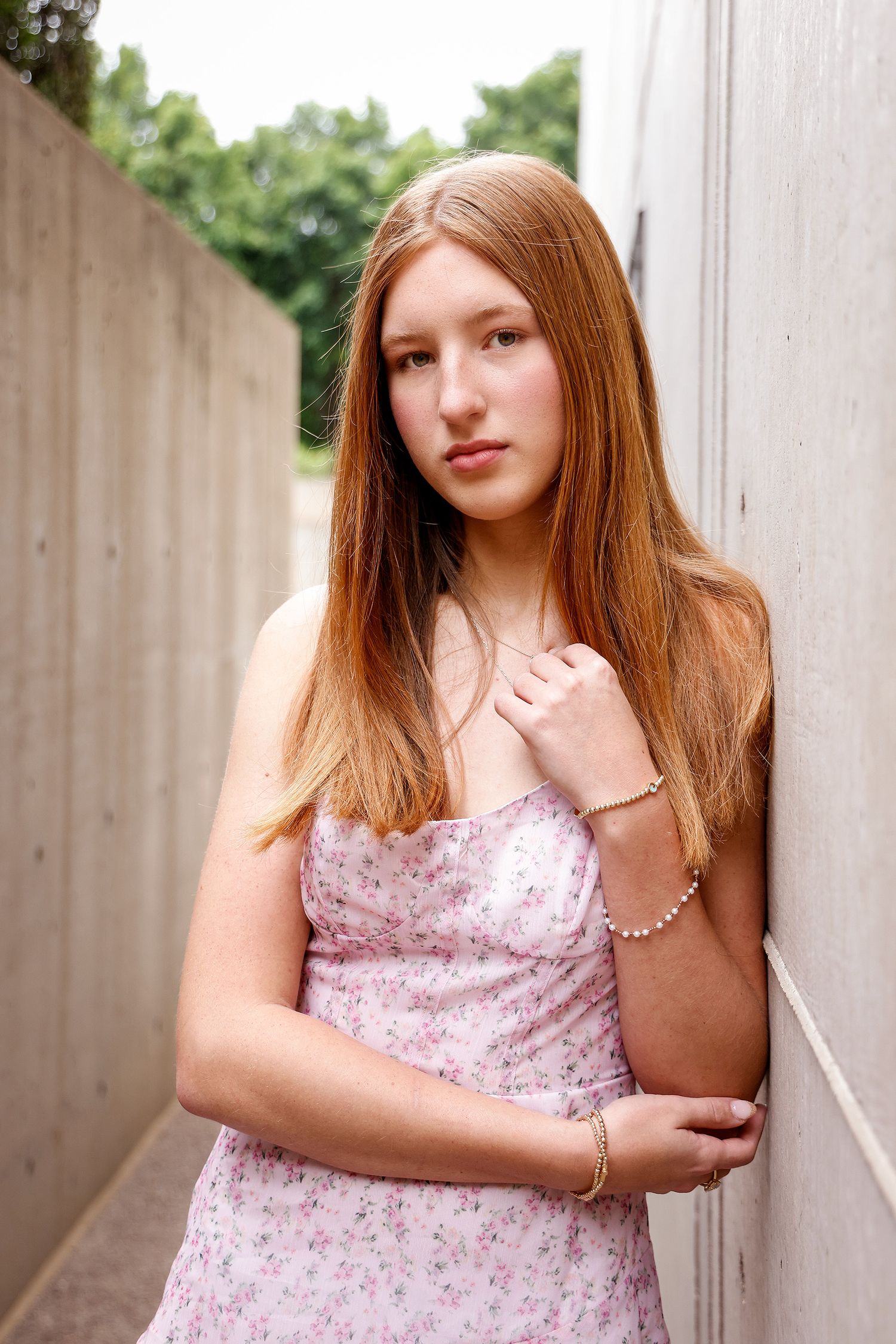 Young woman with long red hair in a pink floral sundress leaning against a concrete wall outdoors.