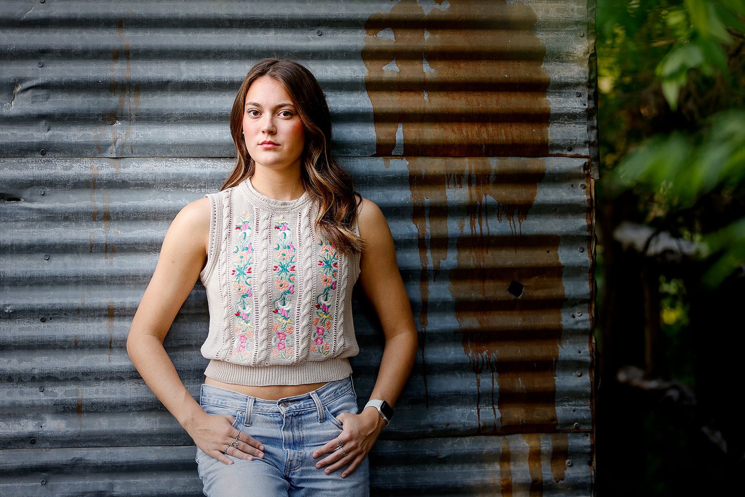Young woman in embroidered crop top and jeans posing confidently against rustic corrugated metal wall outdoors.