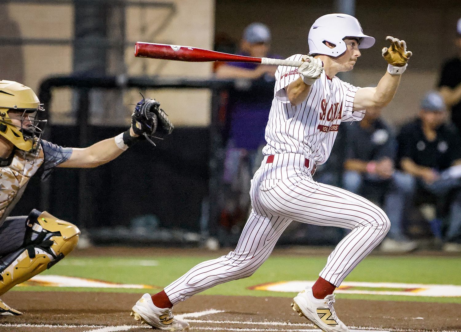 College baseball player in red pinstripe uniform swings bat during night game, catcher and umpire visible behind.