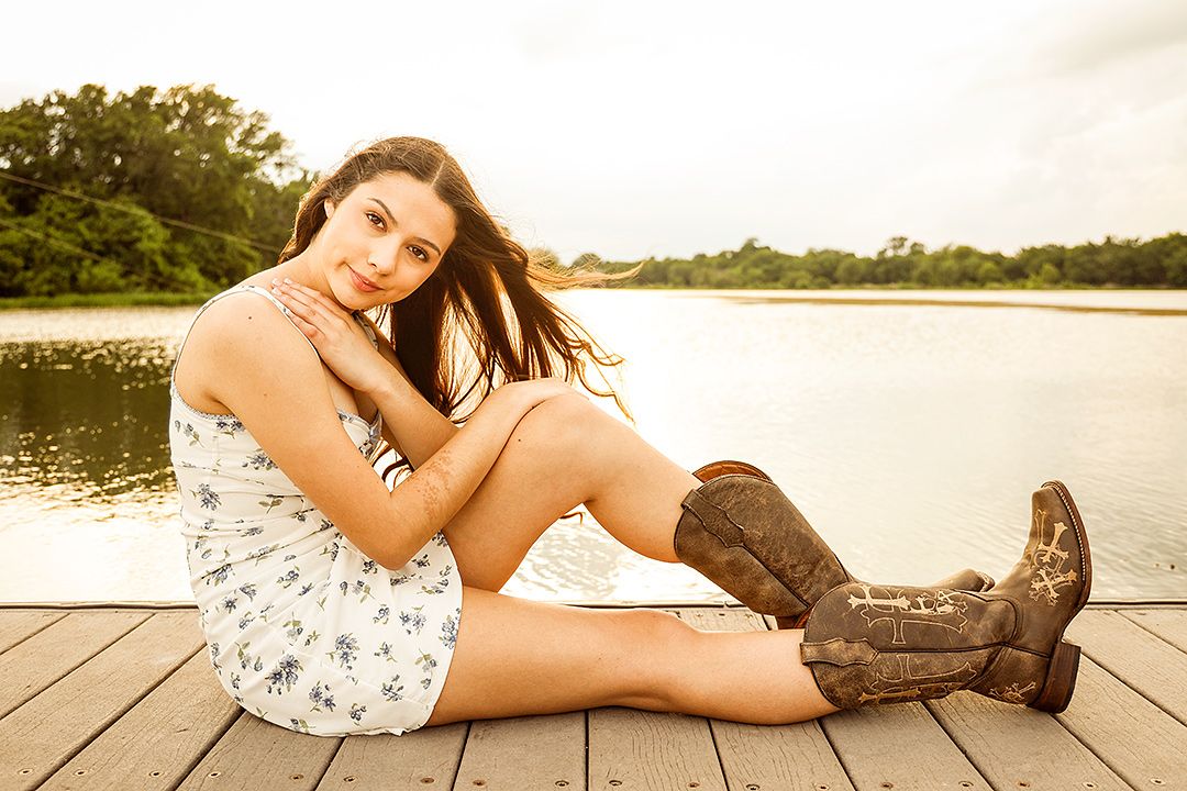 Young woman in floral dress and cowboy boots sitting on a wooden dock by a lake on a sunny day.