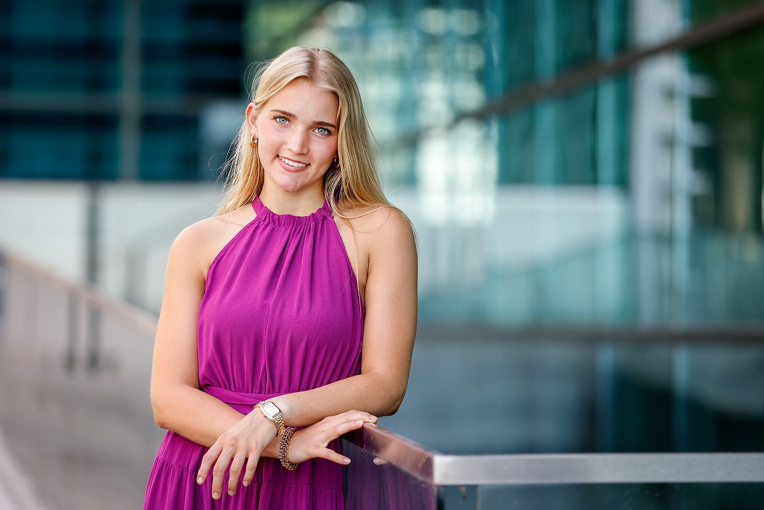 Young blonde woman in a magenta halter dress smiling outdoors near modern glass building.