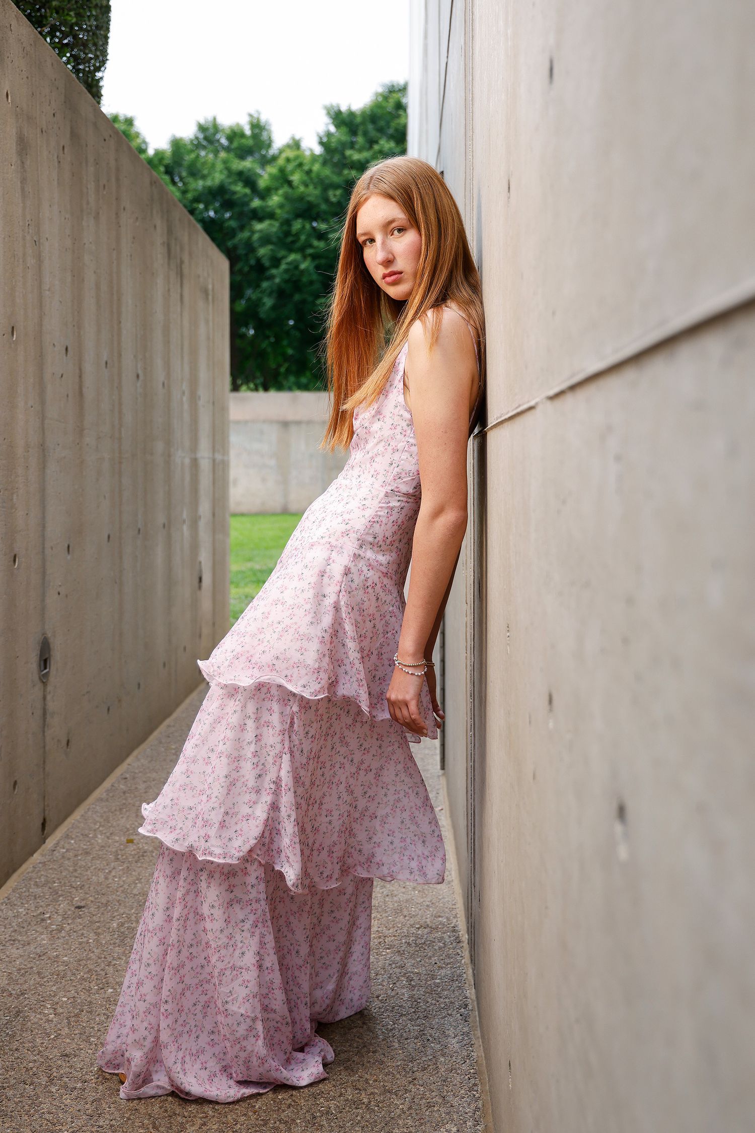 Young woman in a pink floral tiered maxi dress leaning against a concrete wall outdoors.