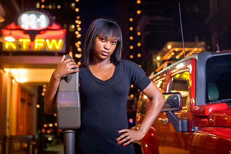 Young Black woman in black outfit poses at night near a parking meter with neon city lights behind her.