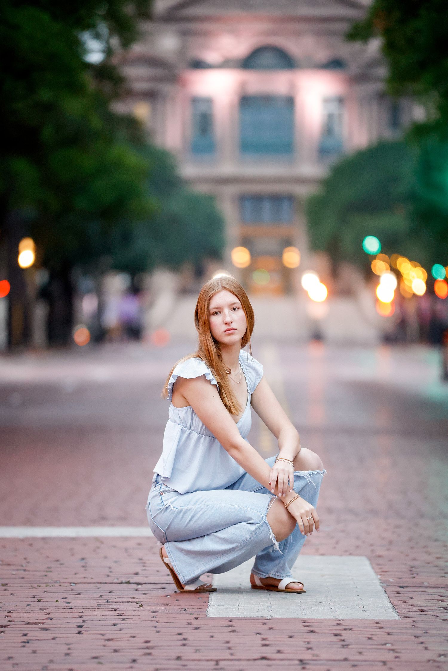 Young woman in blue top and jeans crouching on brick plaza at dusk with bokeh city lights behind her.