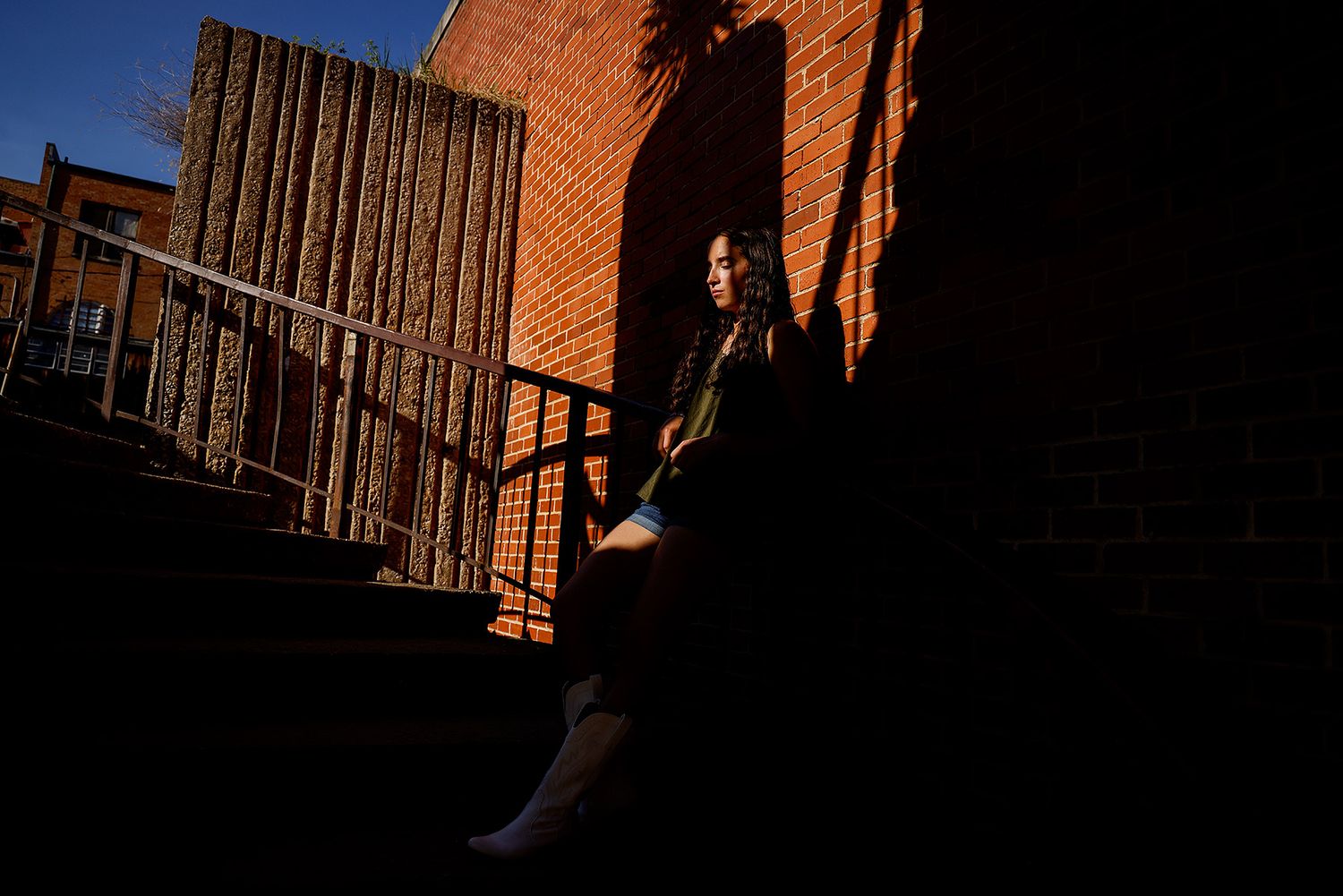 Young woman sitting on sunlit brick steps in dramatic shadow and light, urban outdoor setting.
