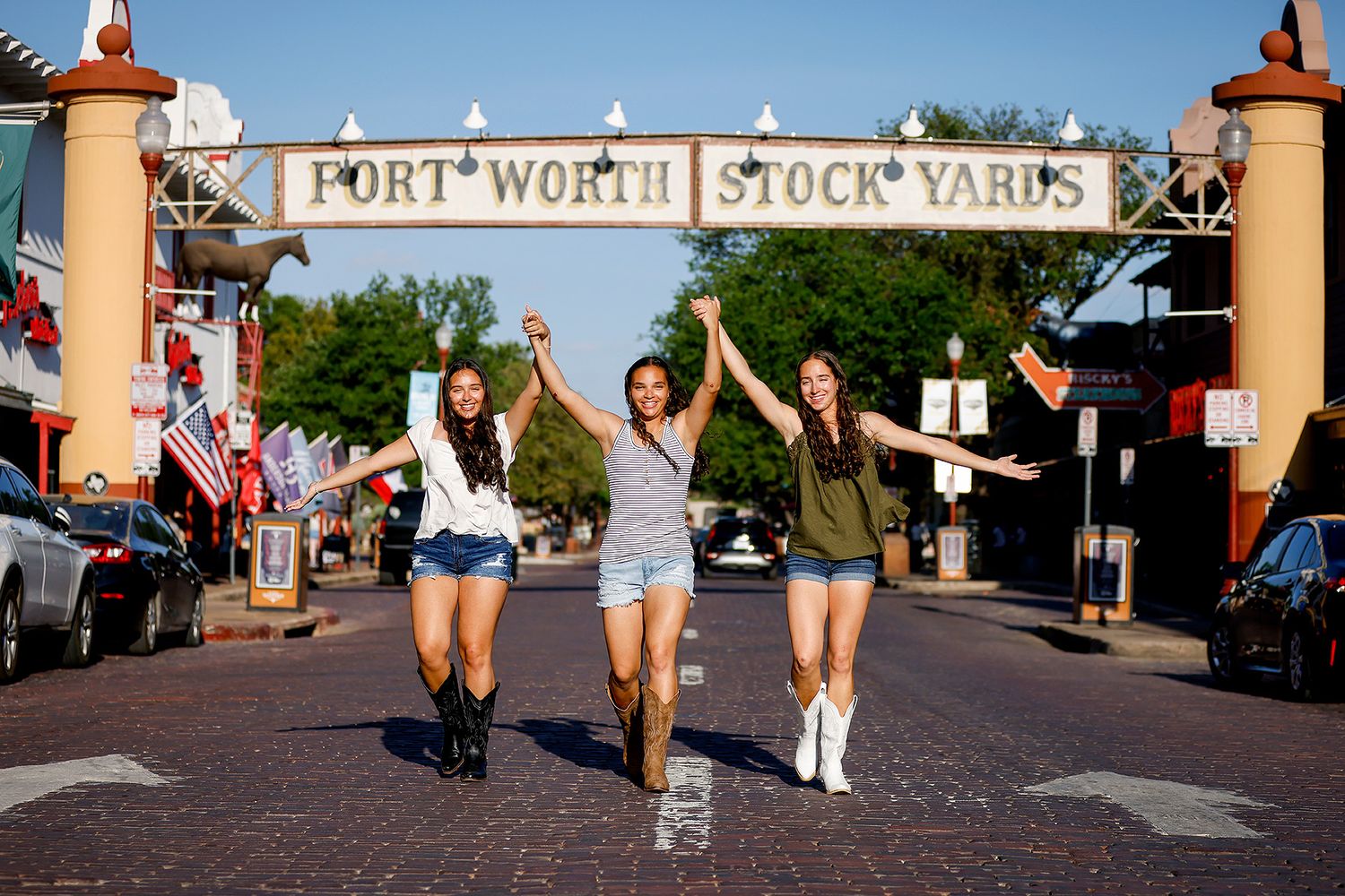 Three women joyfully pose under the Fort Worth Stock Yards sign on a sunny day in Texas.