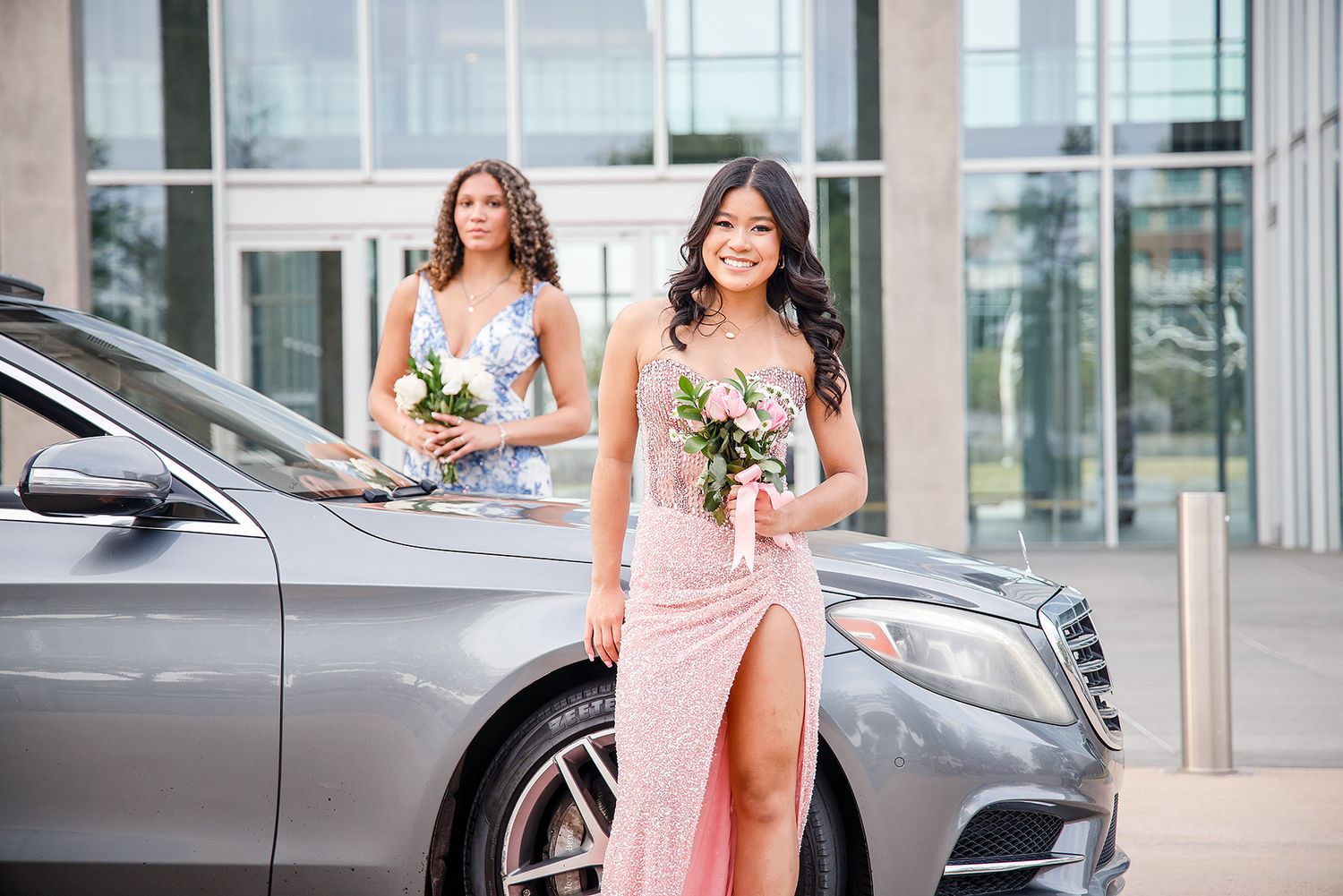 Two young women in elegant prom dresses holding bouquets pose near a luxury Mercedes-Benz car.