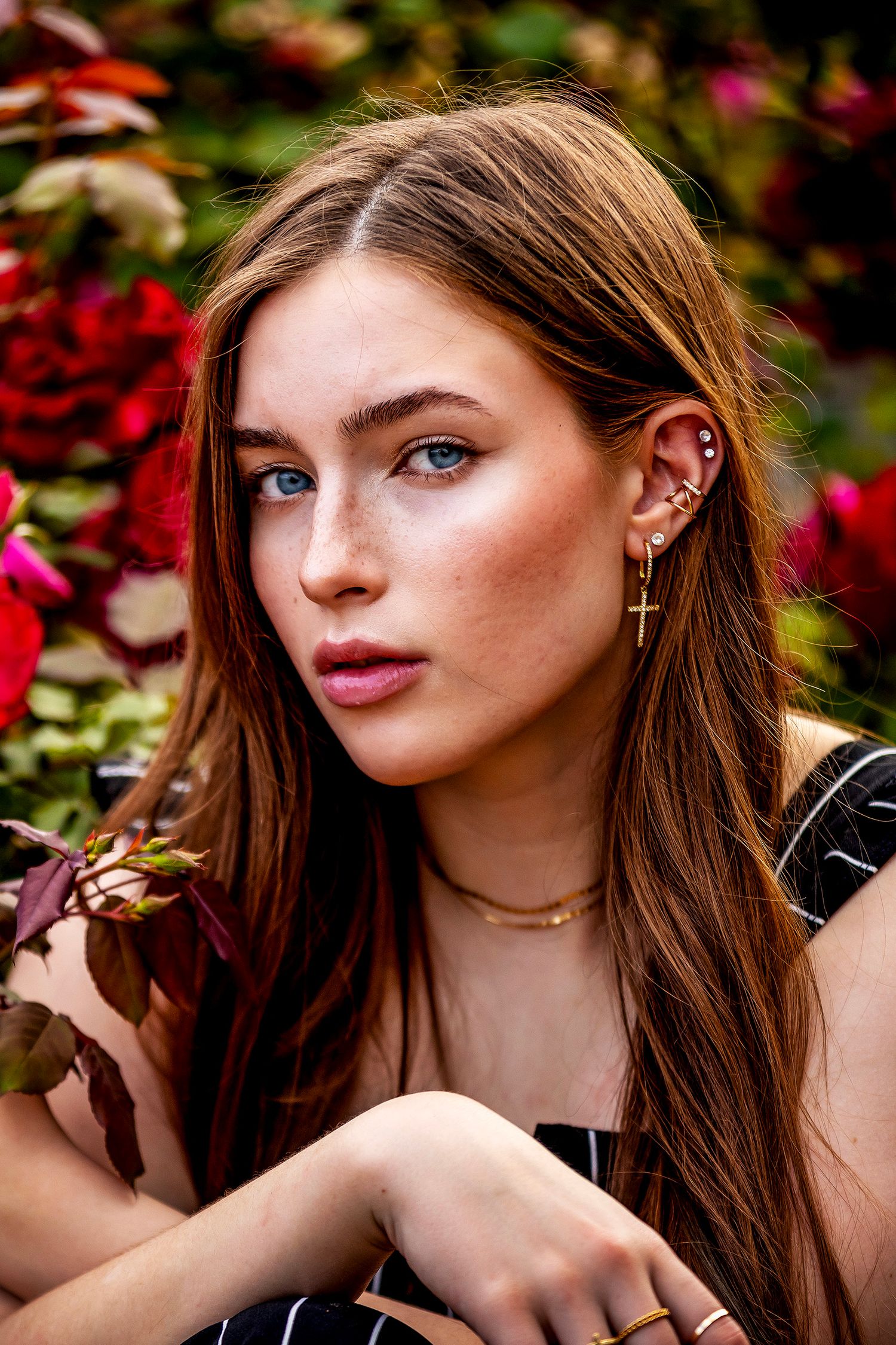 Young woman with long auburn hair and freckles poses among red flowers wearing gold cross earrings and layered necklaces.