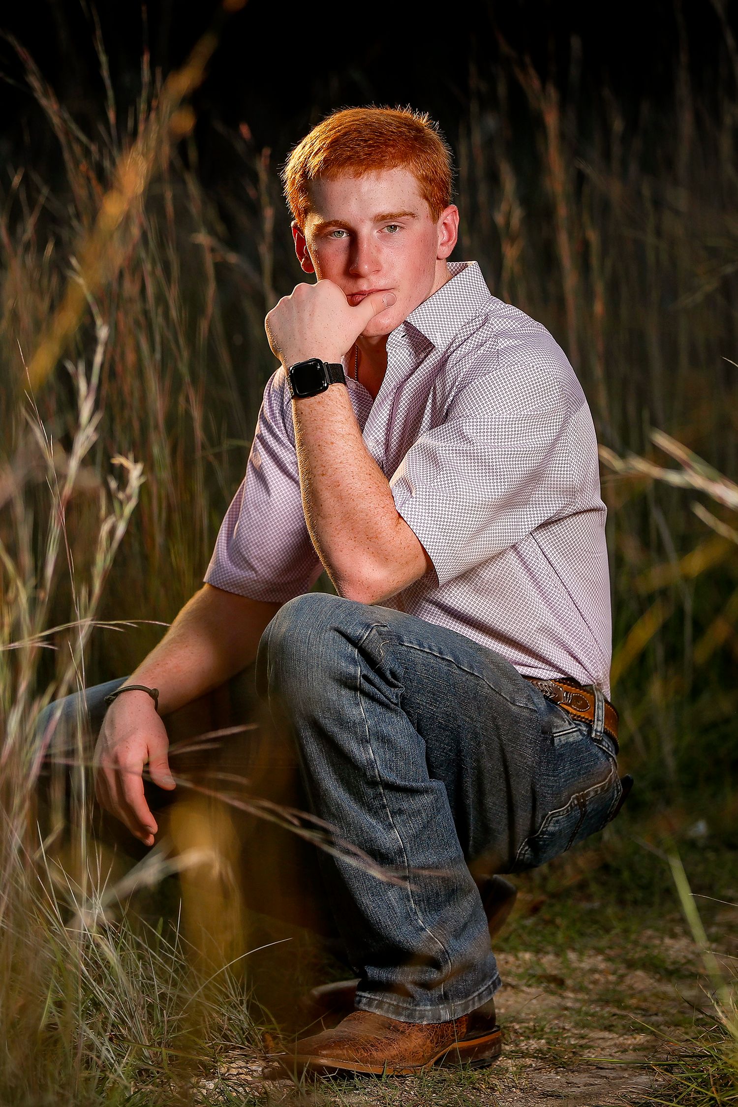 Teenage boy with red hair sits in tall golden grass at dusk wearing jeans and a button-up shirt.