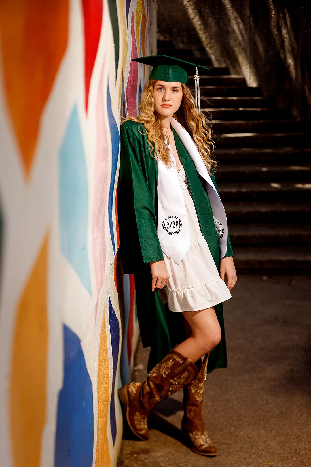 Young woman in green graduation cap and gown with cowboy boots posing by colorful mural wall.