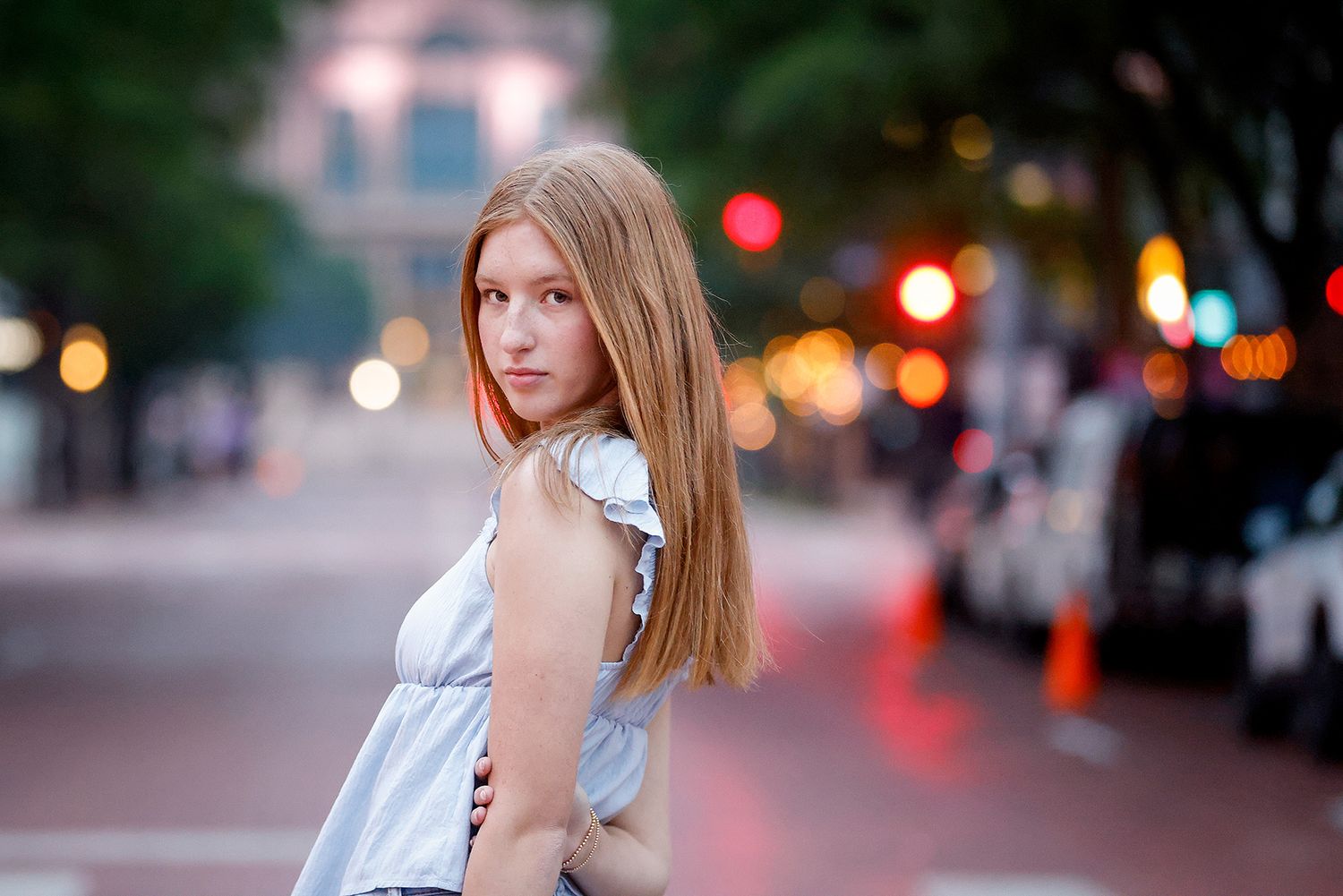 Young woman with long blonde hair in a white top posing on a city street at dusk with bokeh lights.