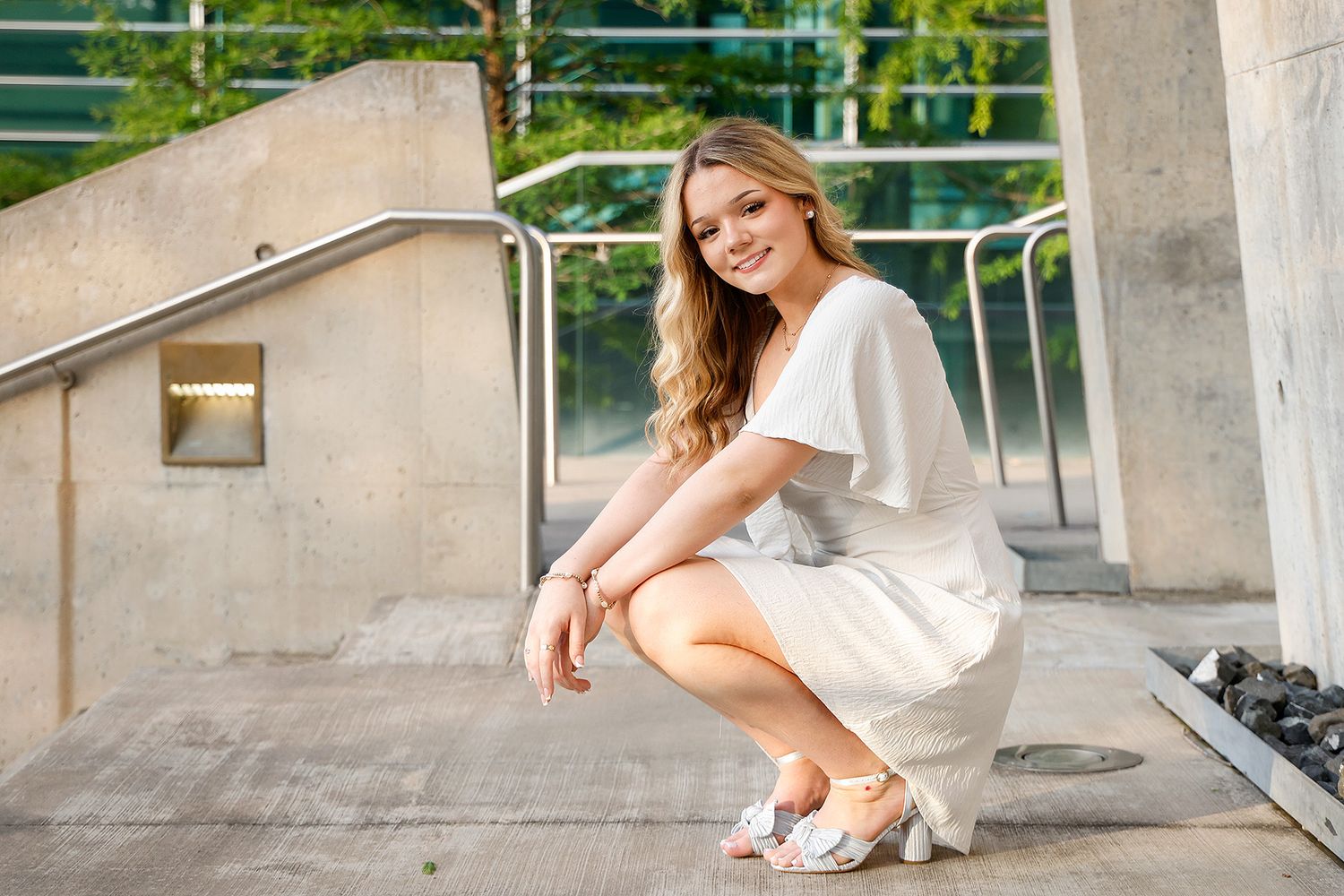 Young woman in white dress and heels crouching gracefully on outdoor steps, smiling confidently in natural light.