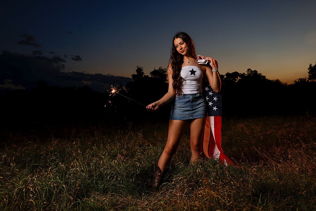 Young woman holding American flag and sparklers in a field at dusk, wearing denim skirt and cowboy boots.