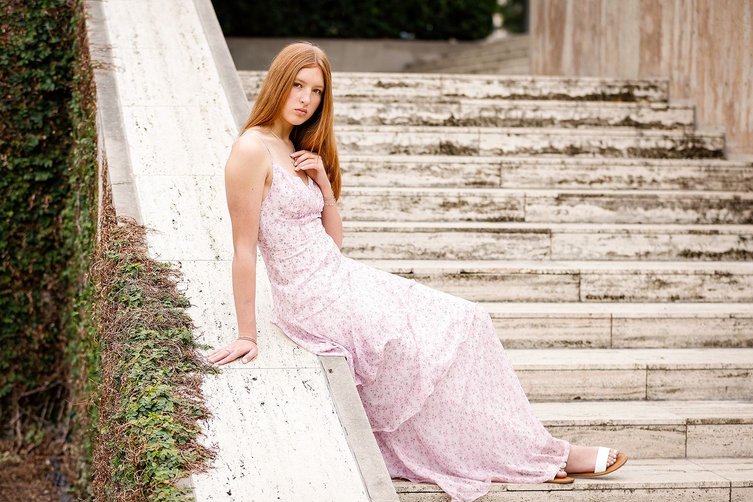 Young woman in a flowing pink floral dress poses gracefully on stone steps outdoors.