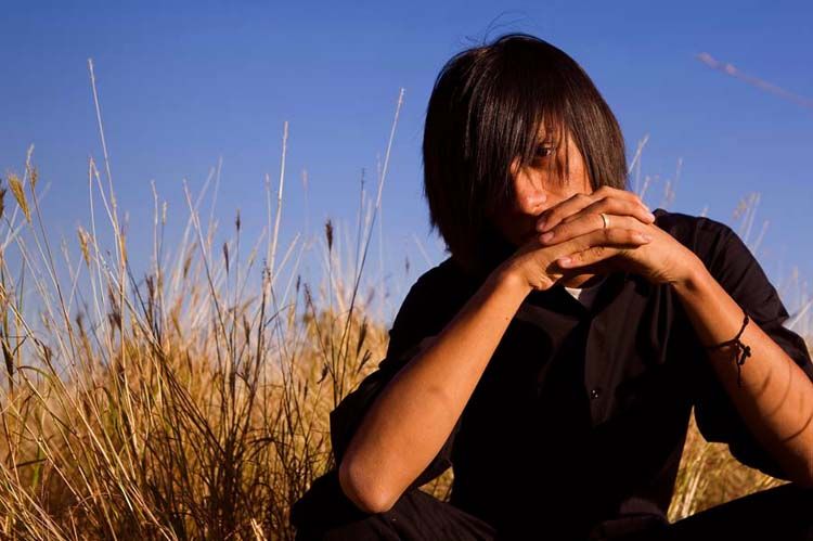 A young person with dark hair sits pensively in a golden wheat field under a clear blue sky.