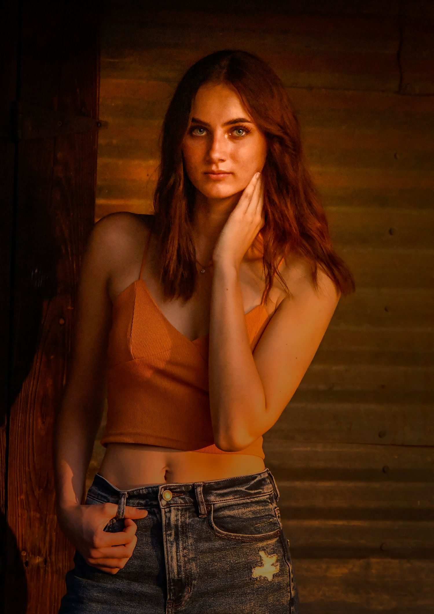 Young woman with auburn hair in orange crop top and jeans poses in warm golden light.