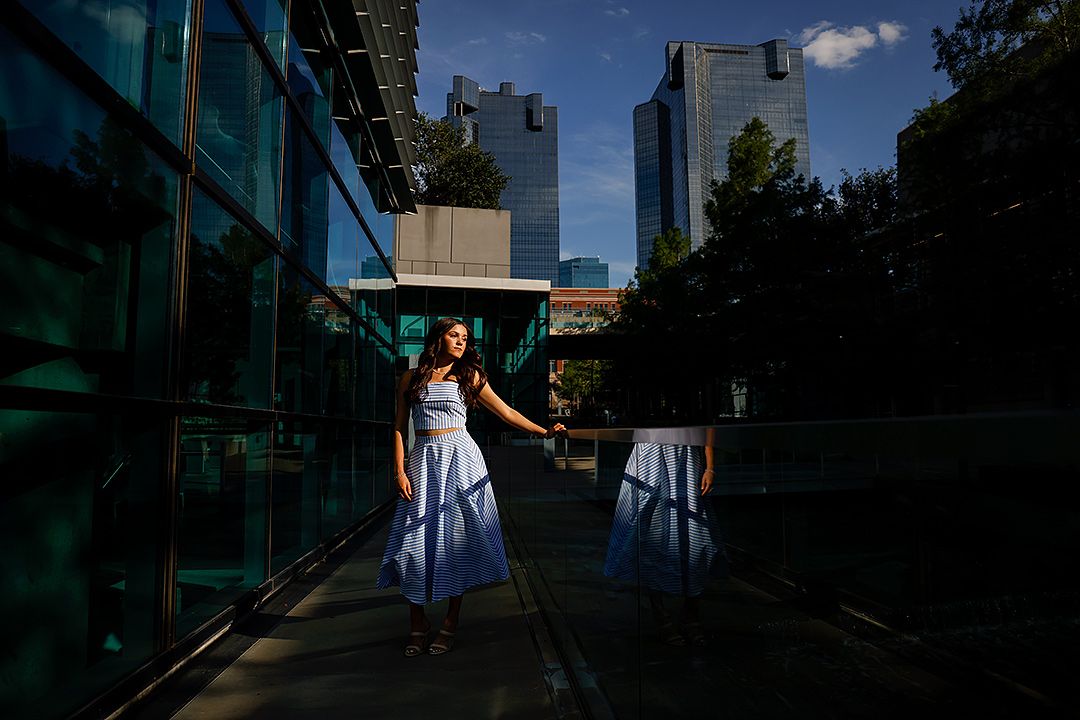 Woman in blue dress poses between glass buildings and skyscrapers on a sunny urban walkway.