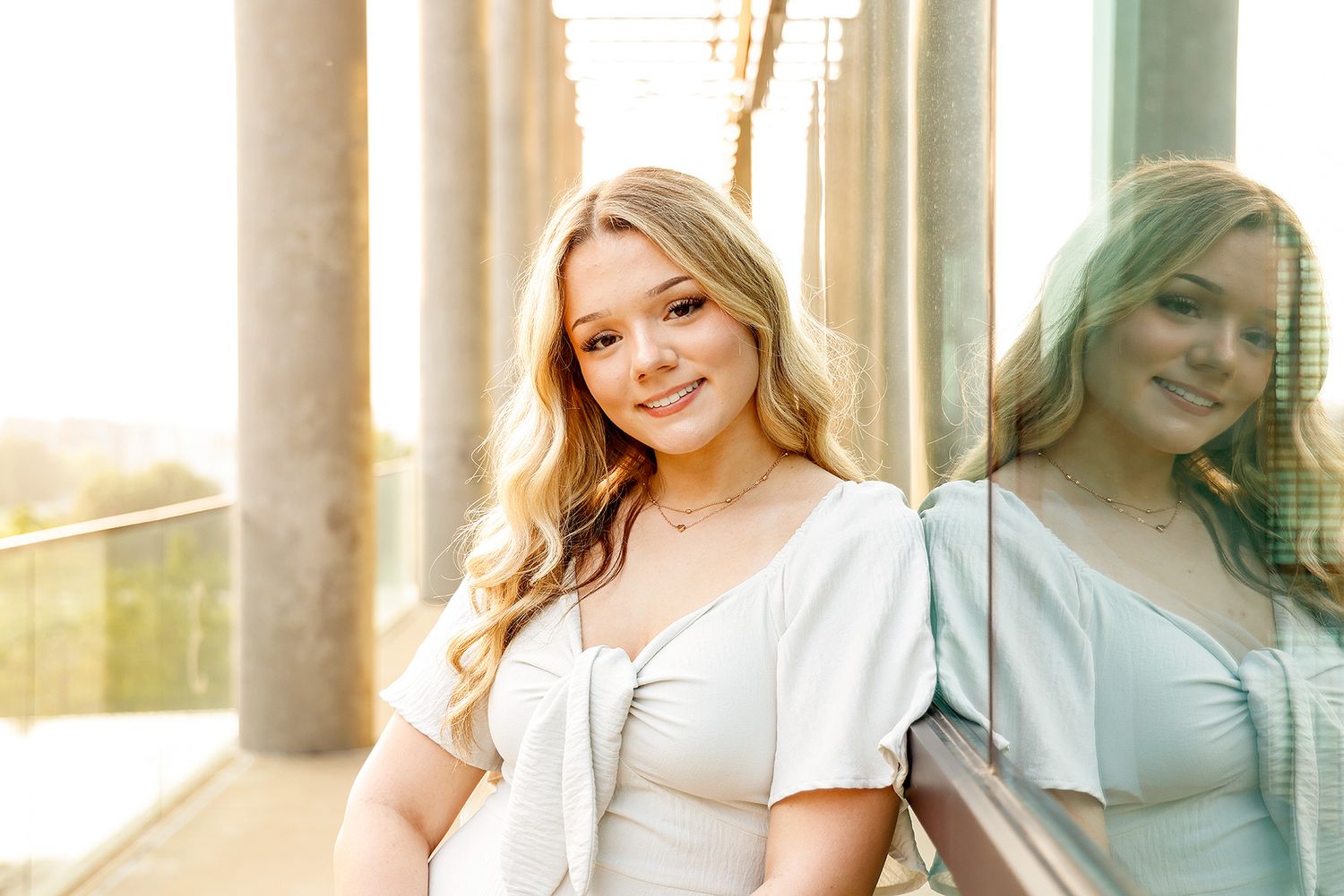 Young blonde woman in white dress smiling, leaning against a reflective glass wall with columns in soft sunlight.