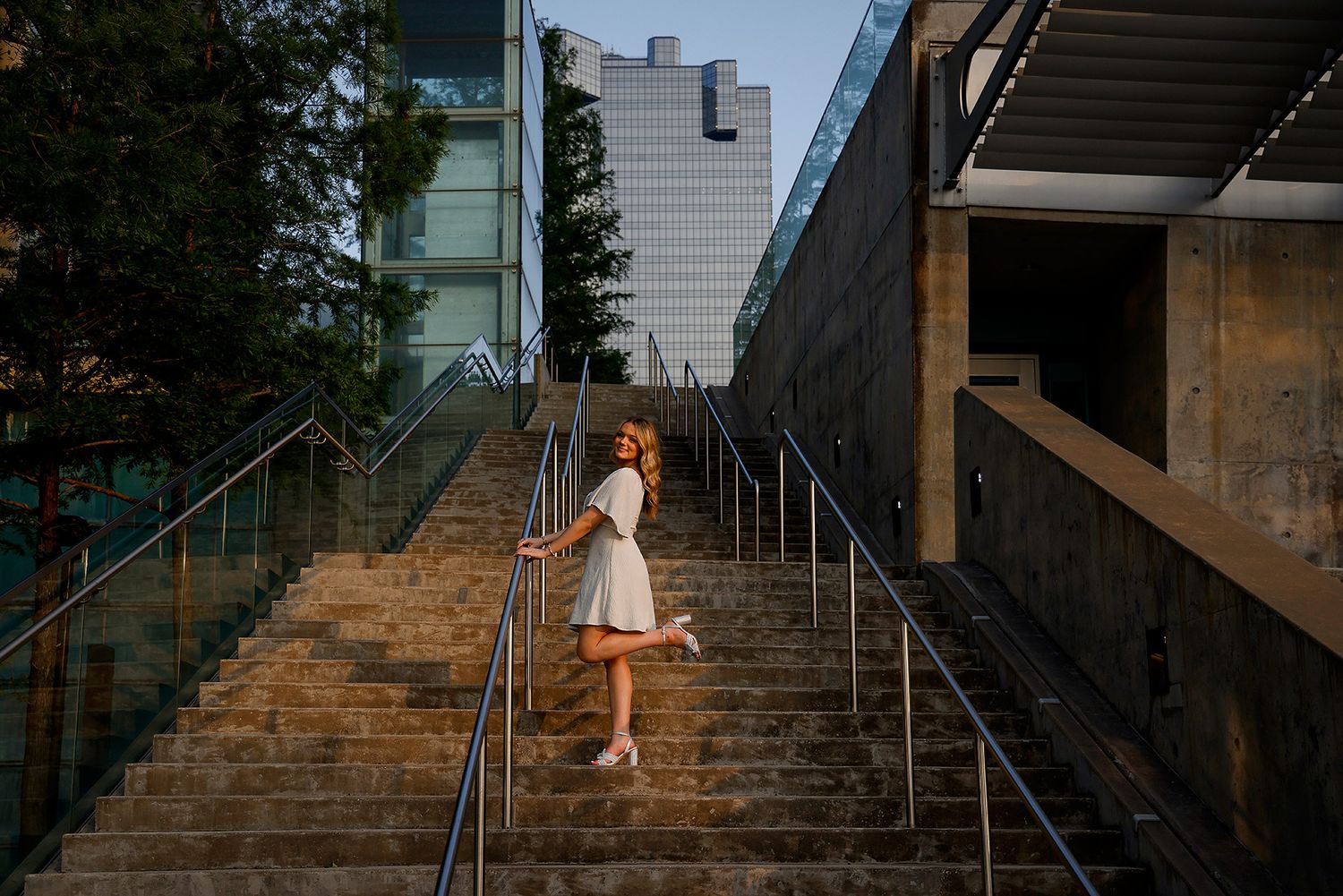 Young woman in white dress posing on urban stairs with modern buildings in background during golden hour.