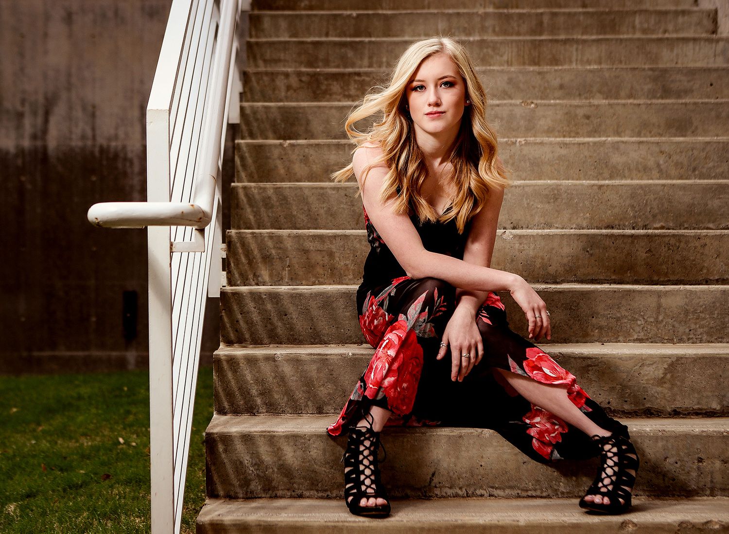 Young blonde woman in floral pants poses on outdoor stone steps with white railing in stylish fashion portrait.