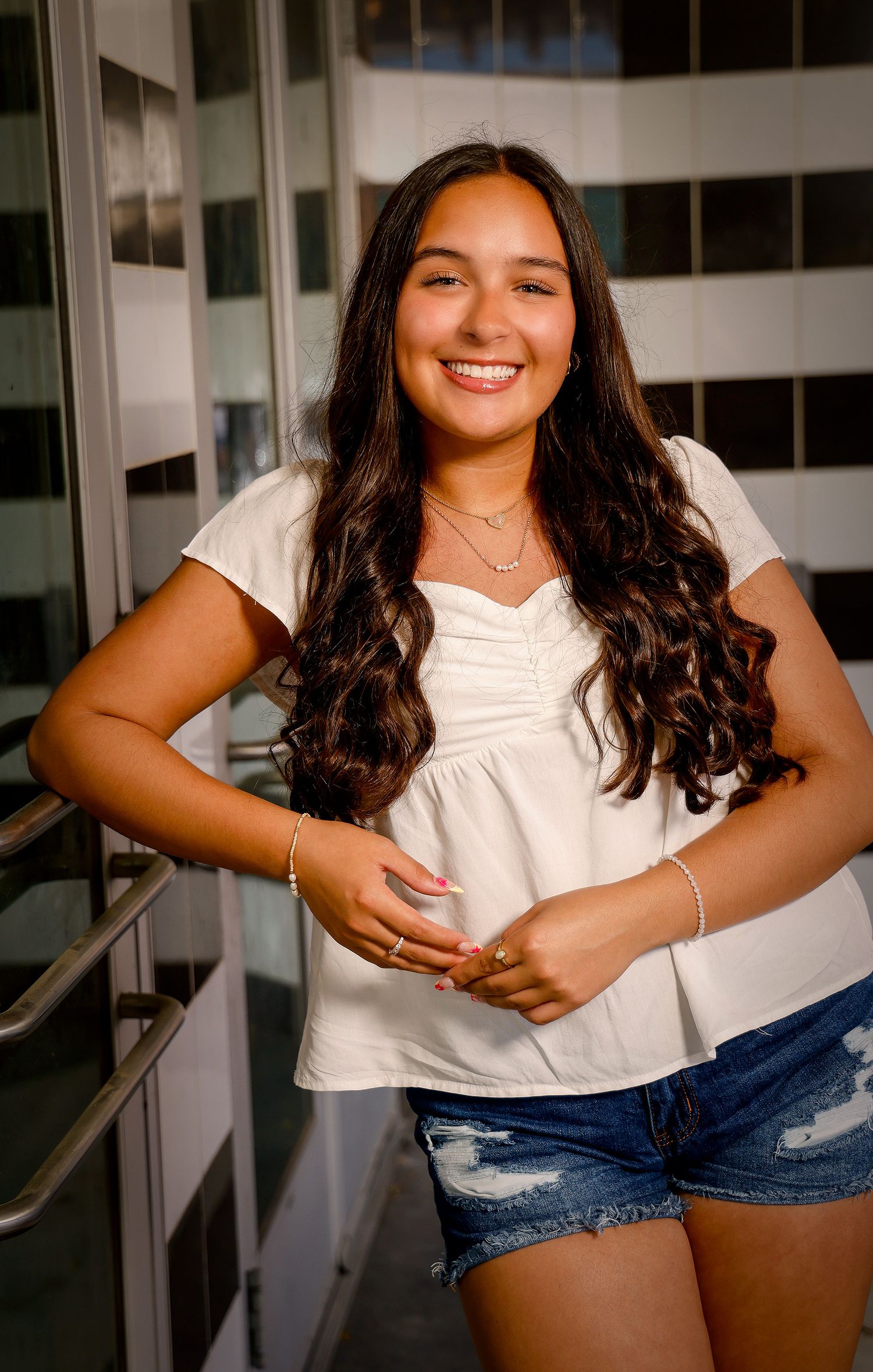 Smiling young woman with long curly hair wearing a white top and denim shorts, posing confidently indoors.