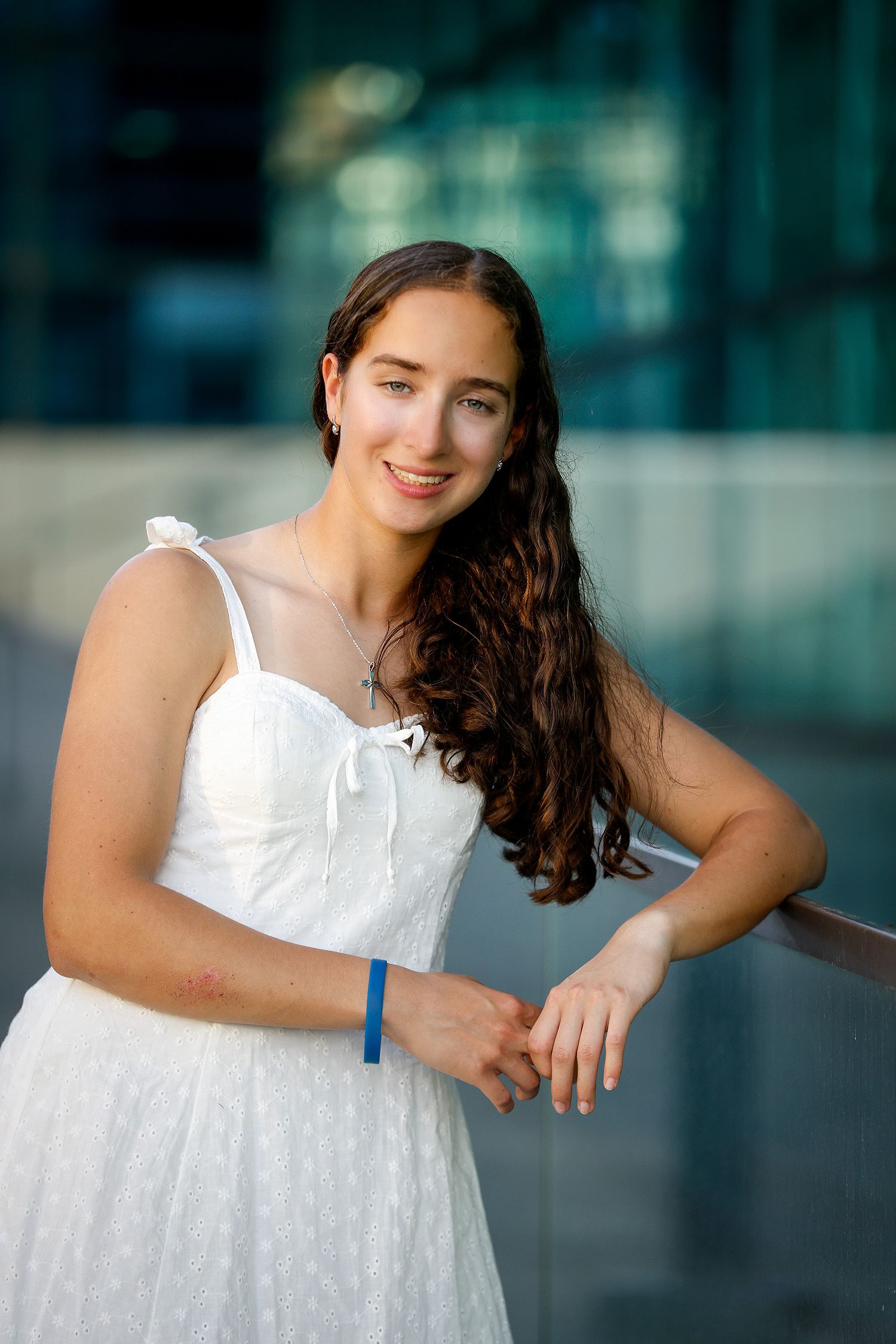 Young woman in white sundress with blue wristband leaning on railing, smiling outdoors near modern glass building.