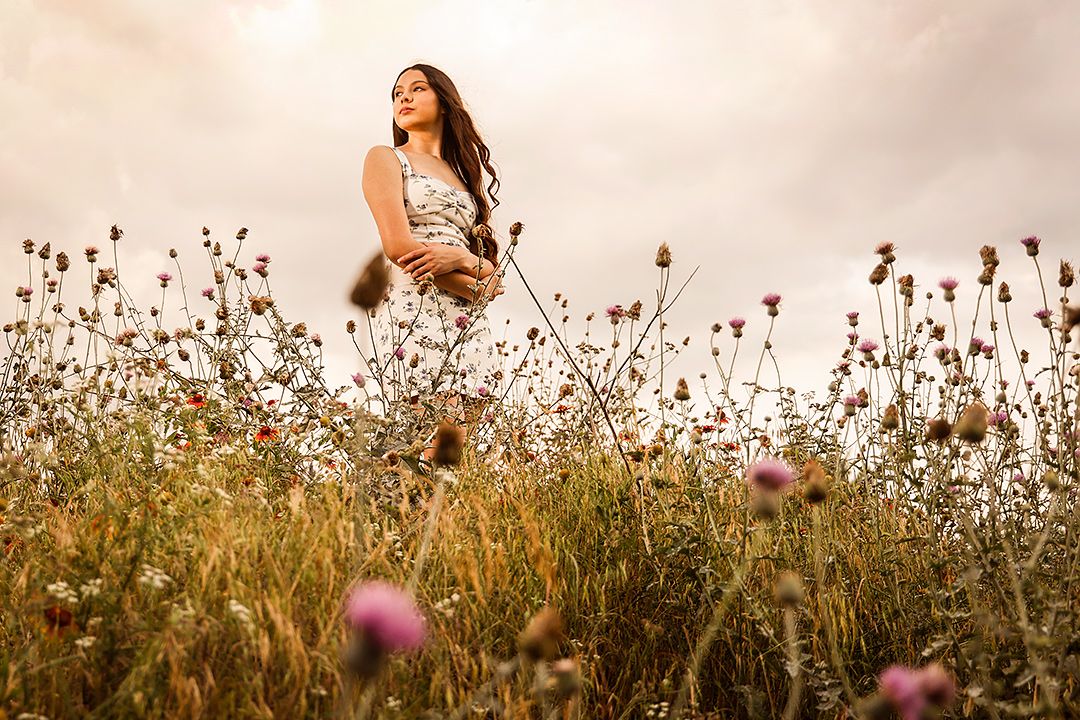 Young woman standing in a wildflower meadow under a dramatic cloudy sky, surrounded by blooming pink and purple flowers.