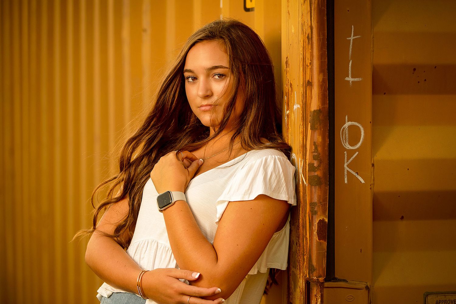 Young woman with long brown hair in white top poses confidently against a golden corrugated metal wall.