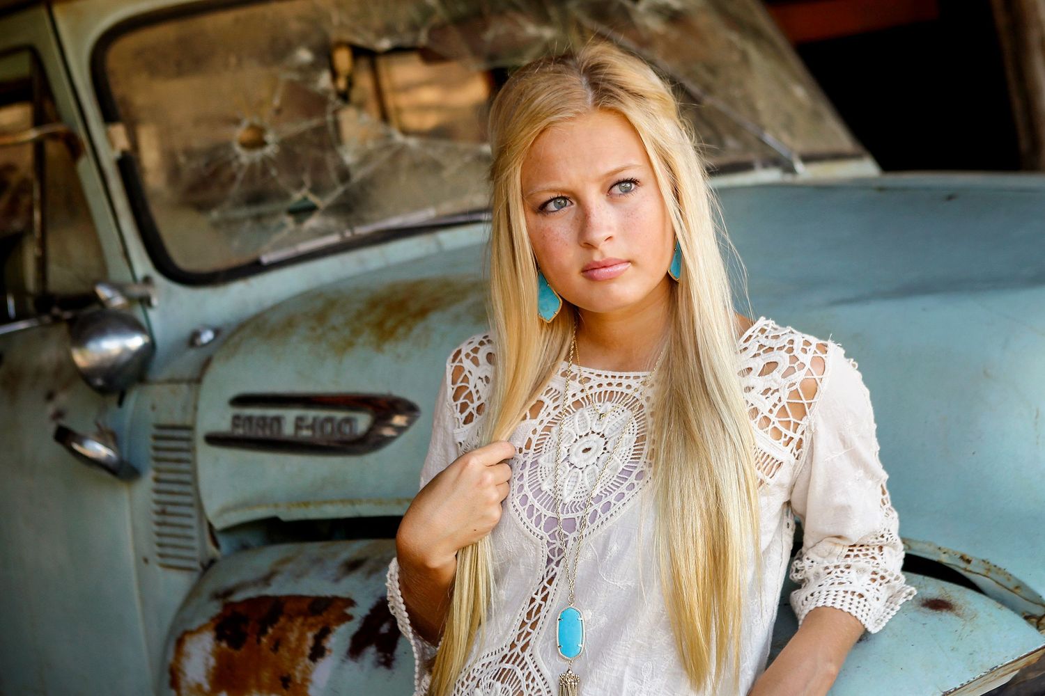 Blonde girl in white lace dress stands before a rustic vintage blue truck in an outdoor setting.