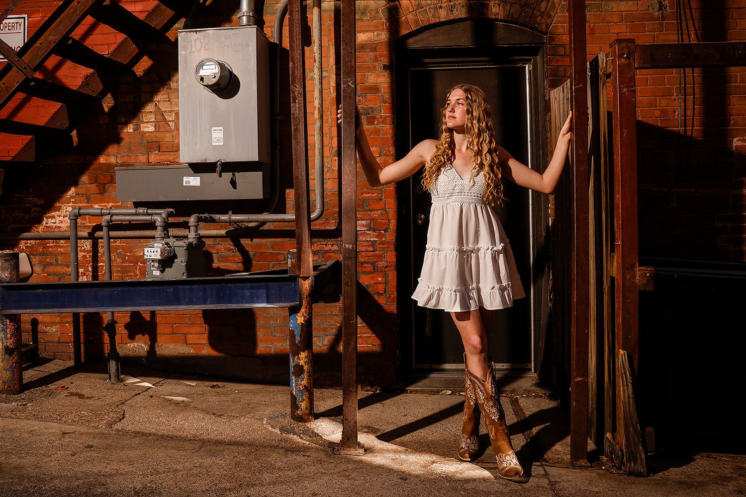 Young woman in white dress and cowboy boots poses dramatically in an urban alley with brick walls and pipes.