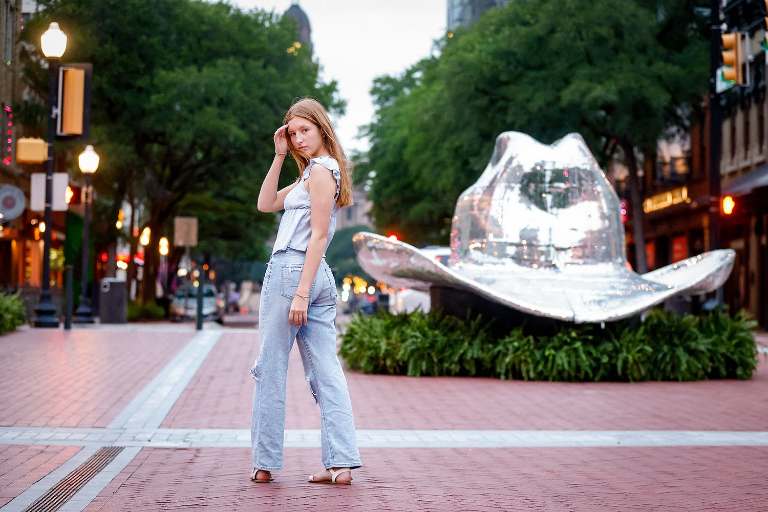 Young woman in jeans poses on a brick plaza beside a large silver cowboy hat sculpture surrounded by trees.