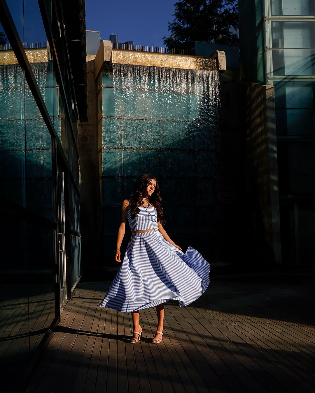 A woman in a light blue two-piece skirt set twirls outdoors near a teal water wall in golden sunlight.