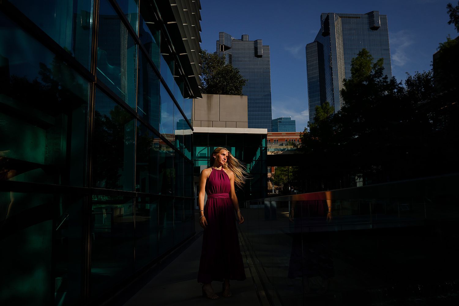 Woman in magenta dress stands confidently in urban setting with glass buildings and trees under dramatic lighting.