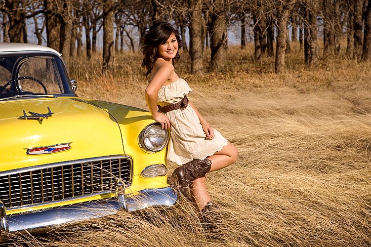 Woman in white dress poses with one leg raised against a vintage yellow classic car in a field.