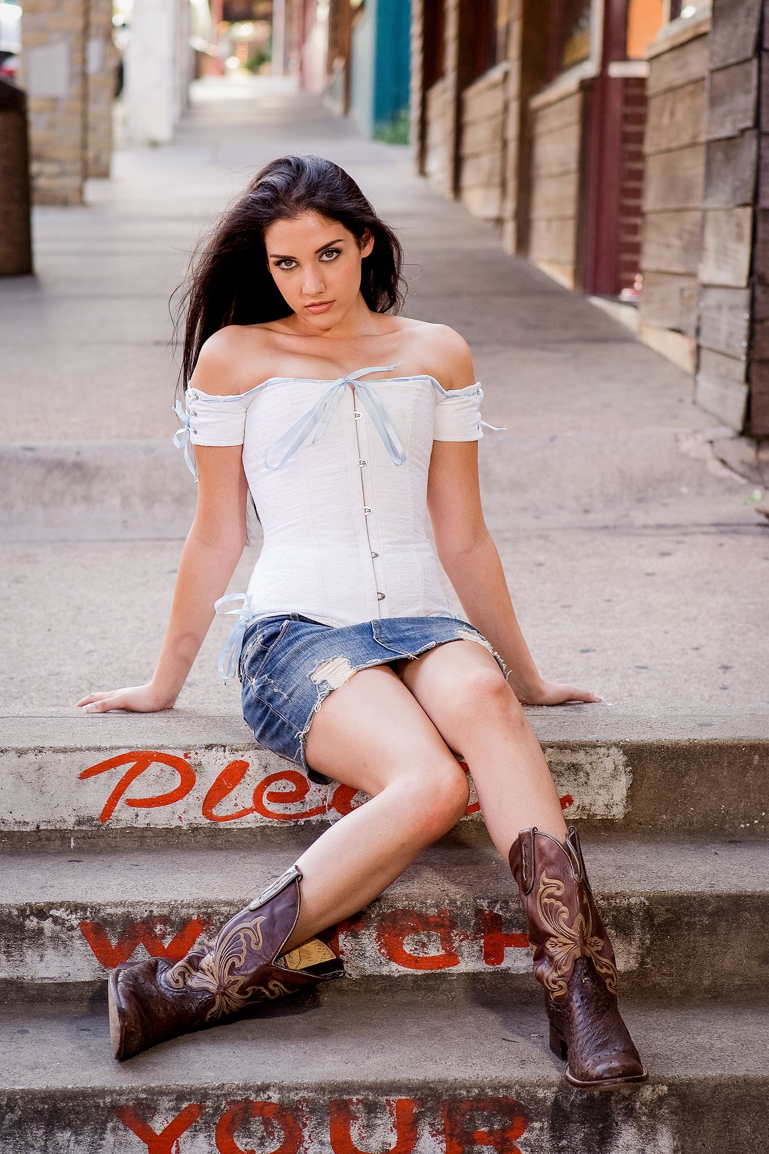 Brunette woman in white corset top and denim skirt sits on steps wearing cowboy boots outdoors.