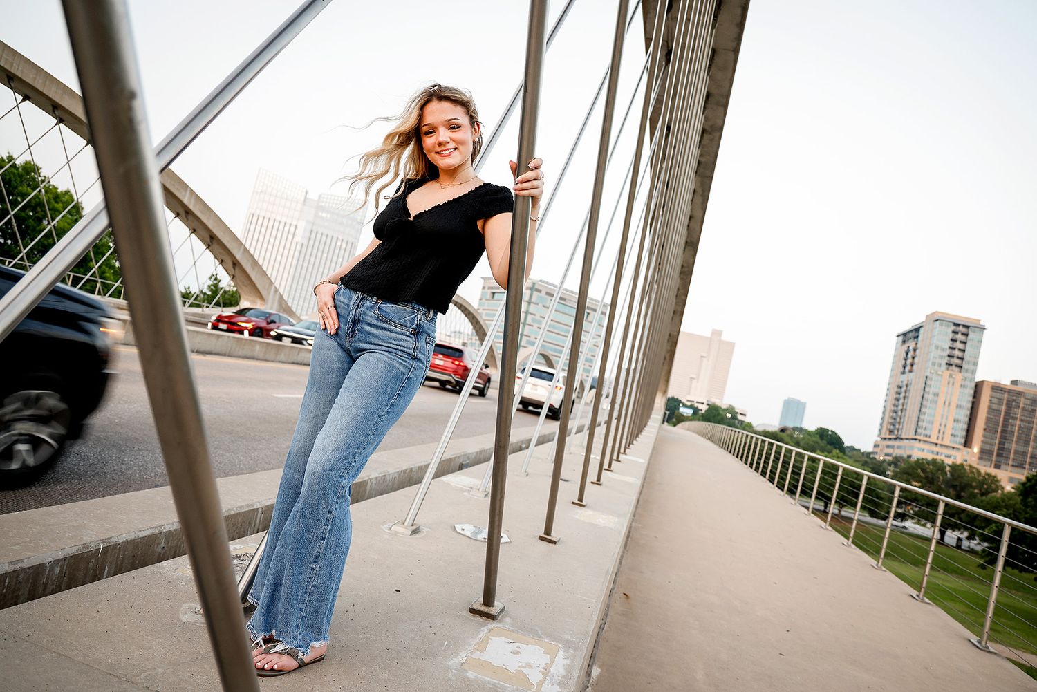 Young woman in black top and flare jeans smiling on a city bridge with urban skyline in background.