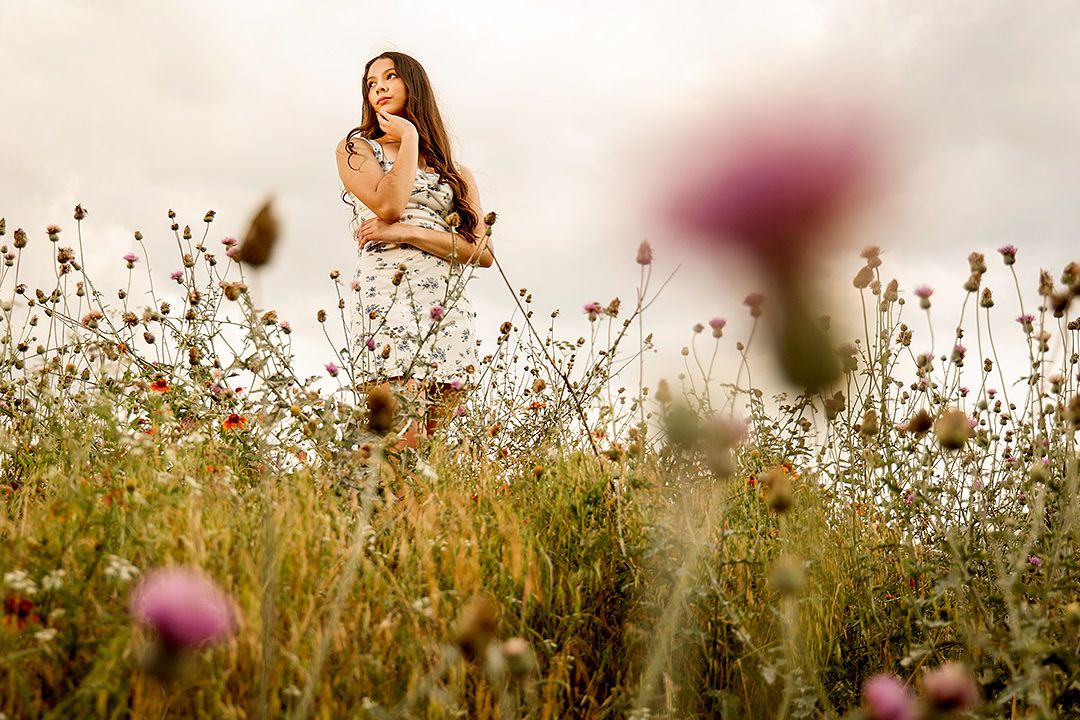 Young woman in white dress standing in a wildflower meadow with pink blooms in foreground under cloudy sky.