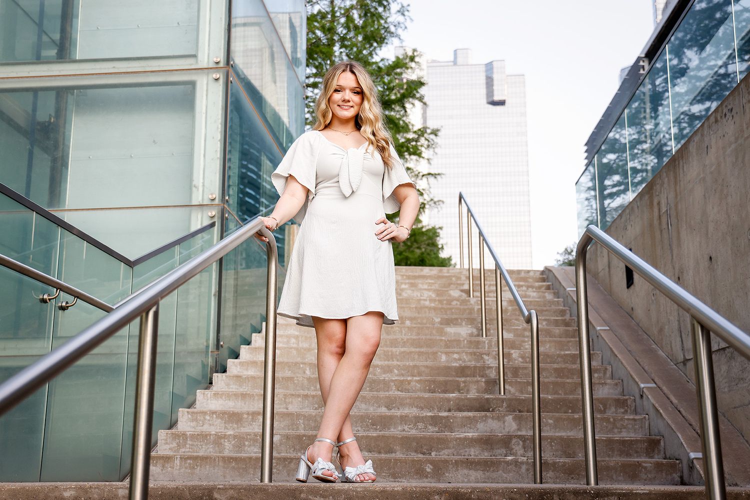 Blonde woman in white mini dress and floral heels posing confidently on outdoor stairs near modern glass building.
