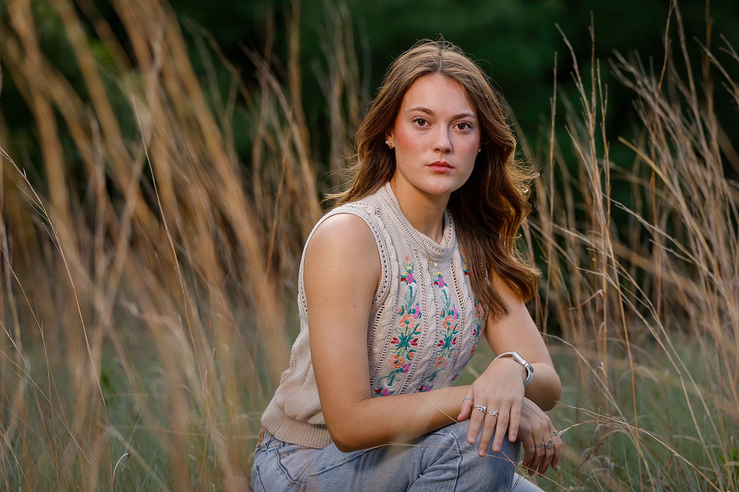 Young woman with long brown hair sits among tall golden grasses in a field, wearing a floral embroidered top and jeans.