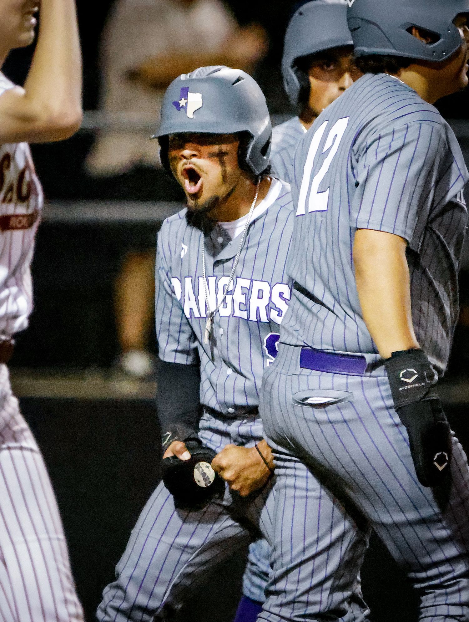 Baseball player in Rangers uniform celebrates enthusiastically on field, pumping fist with teammate wearing number 12 nearby.