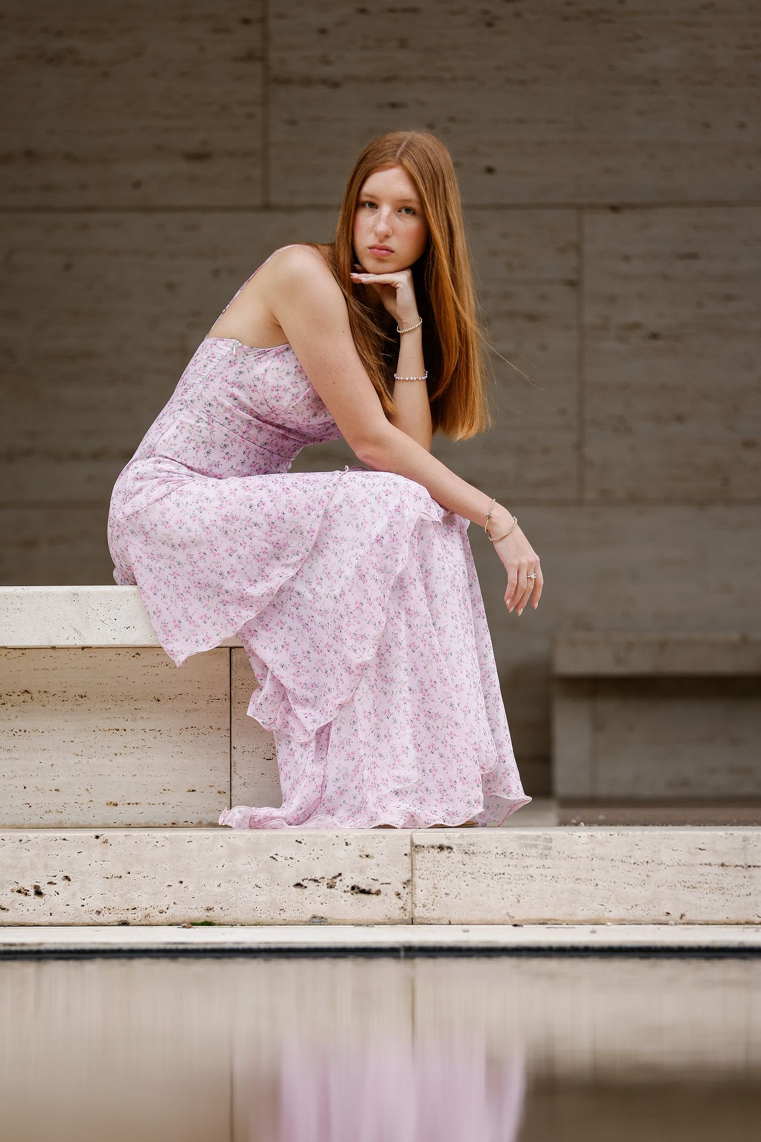 Young woman with red hair in a pink floral strapless maxi dress sitting gracefully on stone steps near water.