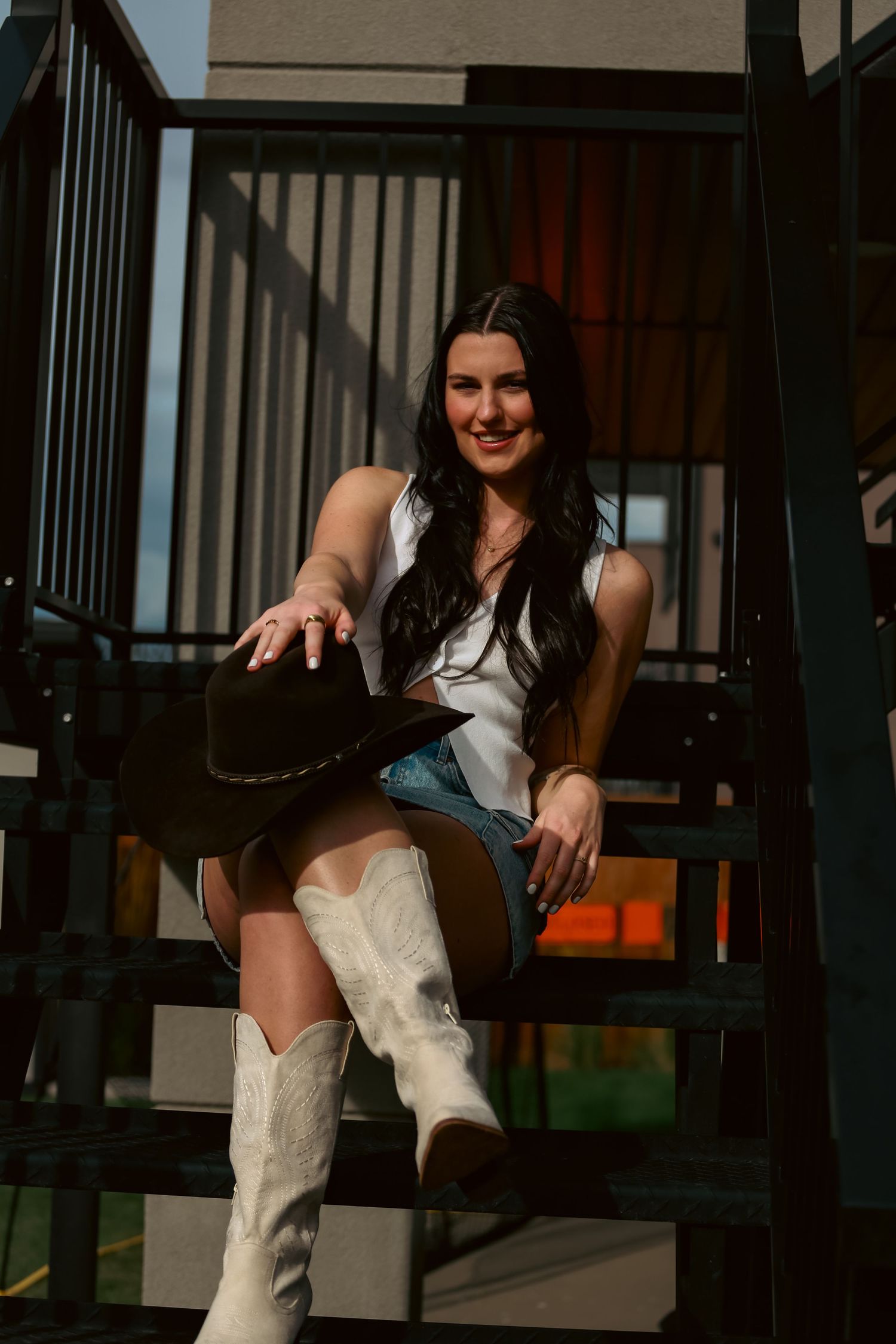 Smiling brunette woman in white top and denim shorts holding black cowboy hat on sunny outdoor staircase.