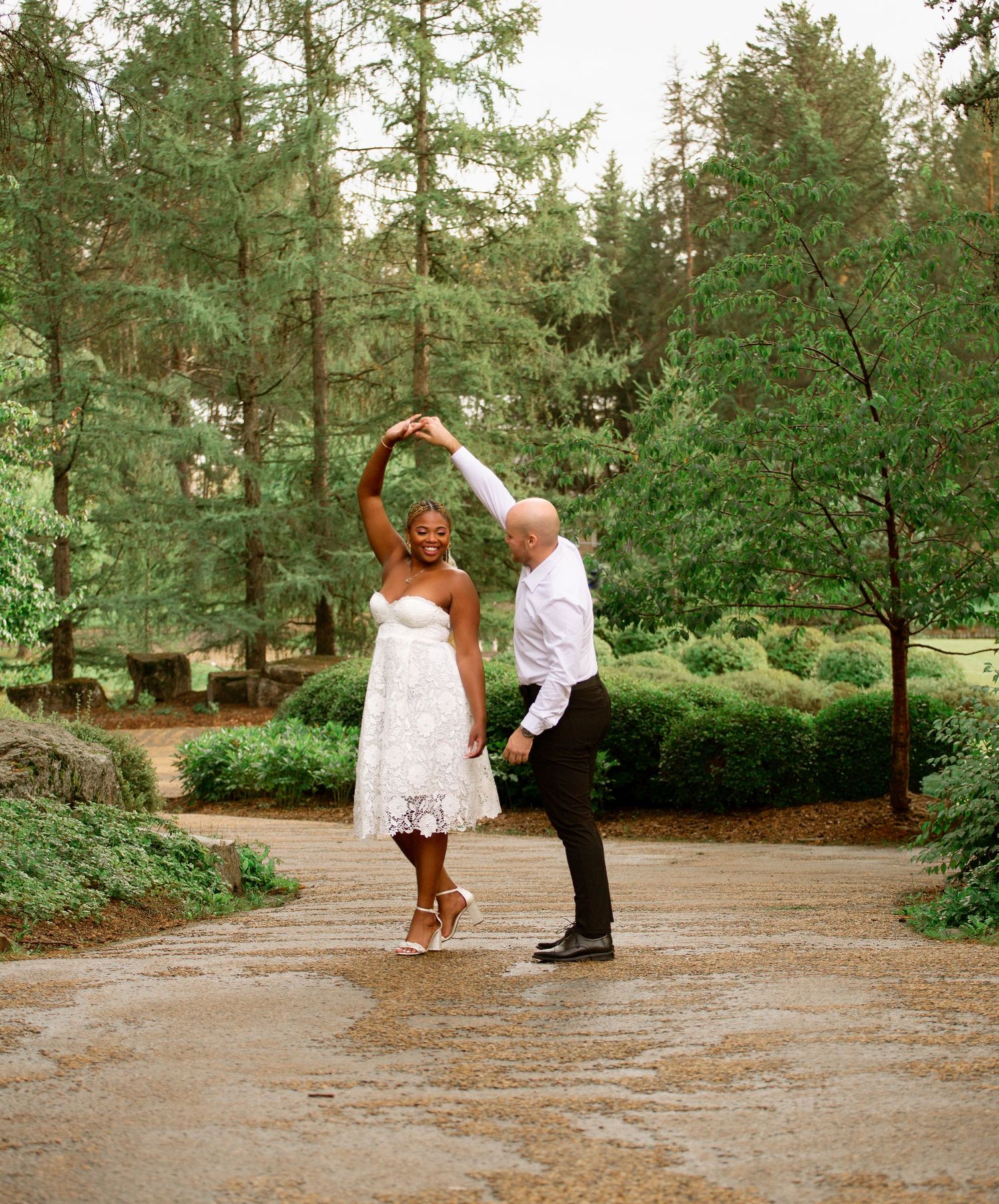 Bride and groom dancing joyfully on a garden path surrounded by tall evergreen trees.