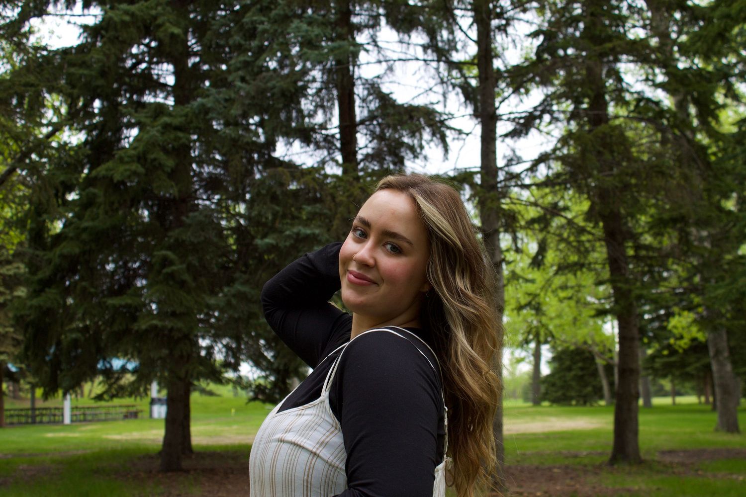Young woman with long blonde hair smiling over shoulder in park surrounded by tall pine trees.