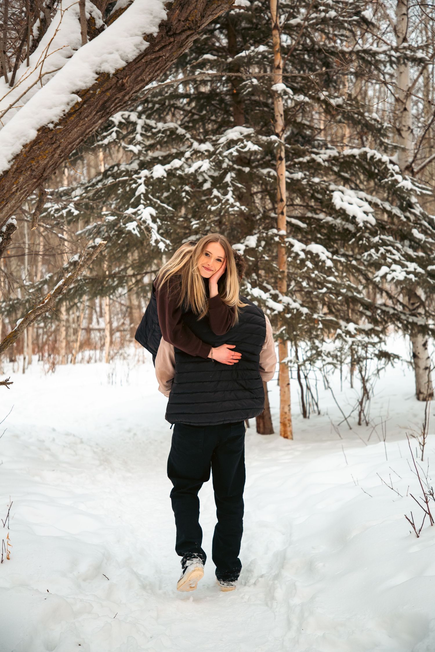 Smiling blonde woman walking through a snowy winter forest during an outdoor engagement photo session.