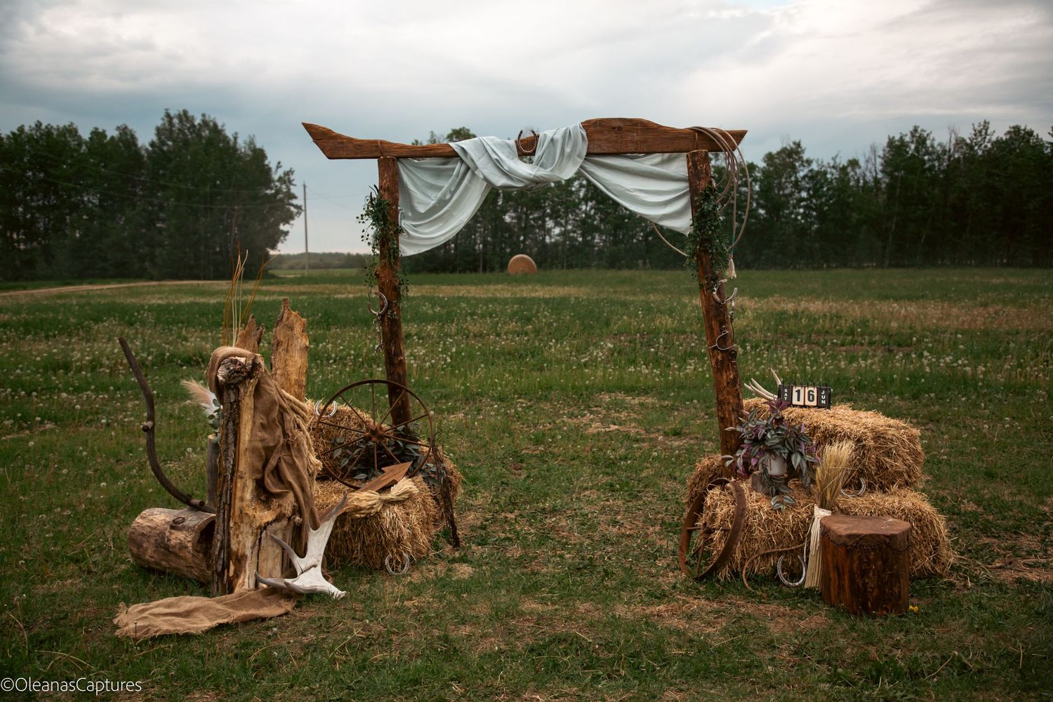 Rustic outdoor wedding arch made of wood and hay bales decorated with white fabric and natural elements in a field.