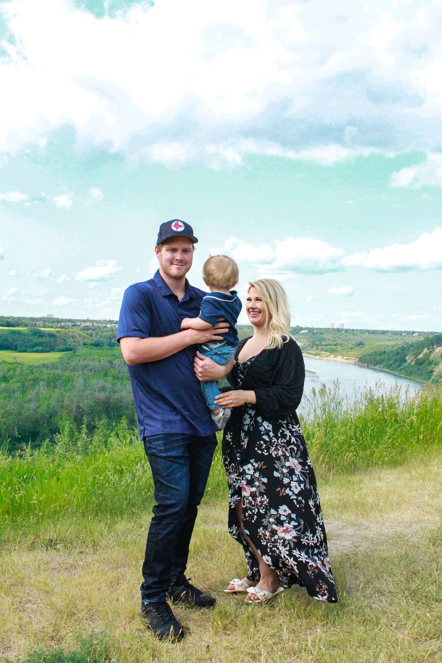 Happy young family with dad holding toddler posing on hilltop overlooking scenic river valley landscape.